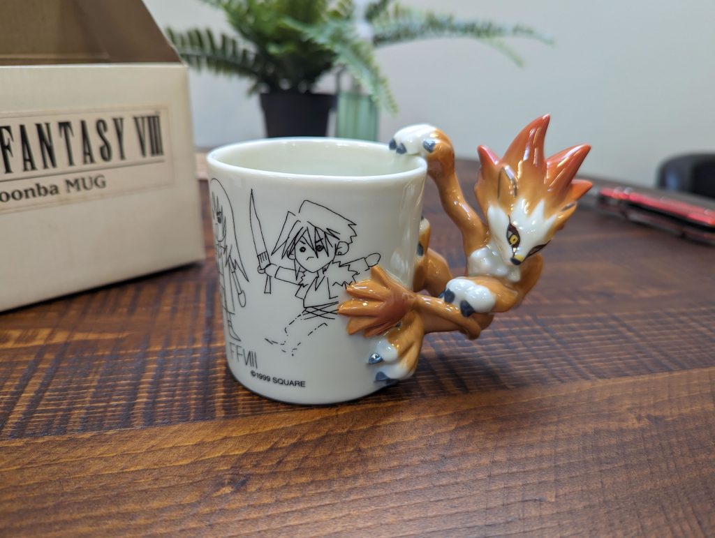 A mug where the handle is a ceramic Moomba figurine clinging to the side. The mug itself is decorated with goofy line drawings of the Final Fantasy VIII cast.